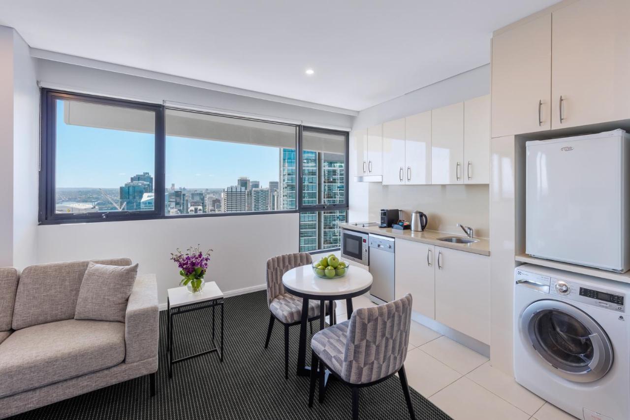 Meriton Suites Kent Street, Sydney Exterior photo The photo shows a modern, well-lit living area that combines a kitchenette and a small dining space. There is a comfortable gray sofa next to a small round table set for two, with a bowl of green apples and a vase of purple flowers on it. The kitchen