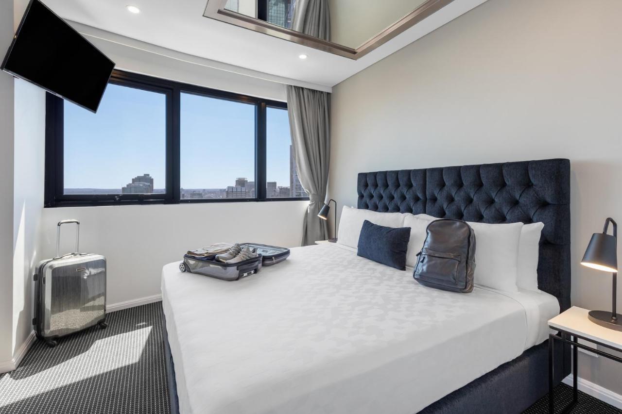 Meriton Suites Kent Street, Sydney Exterior photo The photo shows a modern hotel room featuring a neatly made bed with white linens and a tufted dark headboard. On one side of the bed, there are a couple of bags, likely for travel. The room has large windows with a view of the city, allowing natural