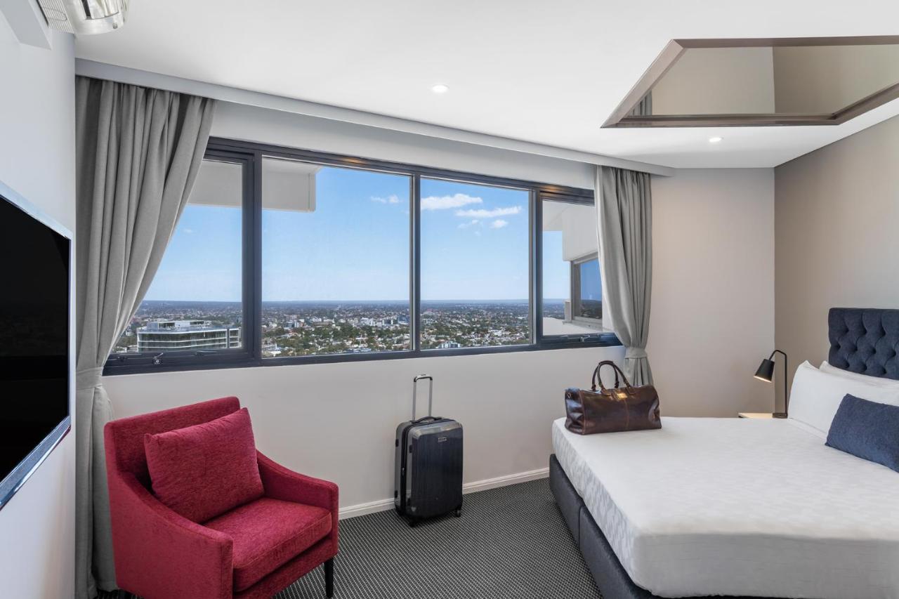 Meriton Suites Kent Street, Sydney Exterior photo The photo shows a modern hotel room. There is a large window with a view, allowing ample natural light to enter. The room features a bed with white bedding, a small red chair, and a black suitcase on the floor. A leather bag is placed on the nightsta