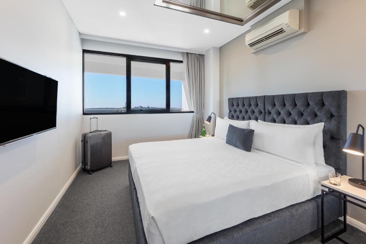 Meriton Suites Kent Street, Sydney Exterior photo The photo shows a modern hotel room with a stylish design. The room features a large, neatly made bed with a tufted gray headboard and white bedding. Next to the bed, there's a small nightstand with a lamp. A sleek flat-screen TV is mounted on the wa
