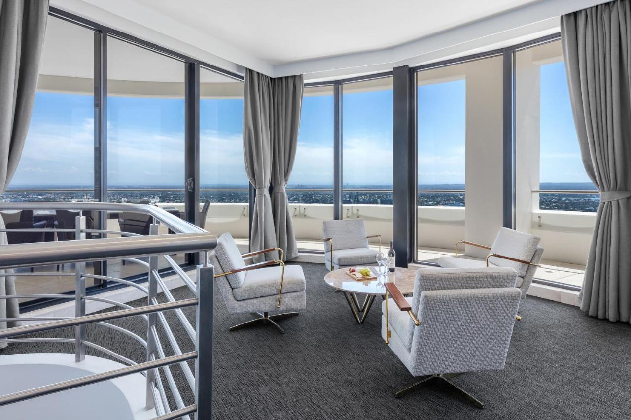 Meriton Suites Kent Street, Sydney Exterior photo The photo shows a modern, stylish living space with large windows that offer a panoramic view of the outdoors. There are several comfortable chairs arranged around a small table, which holds a few items, possibly snacks or drinks. The decor is minima