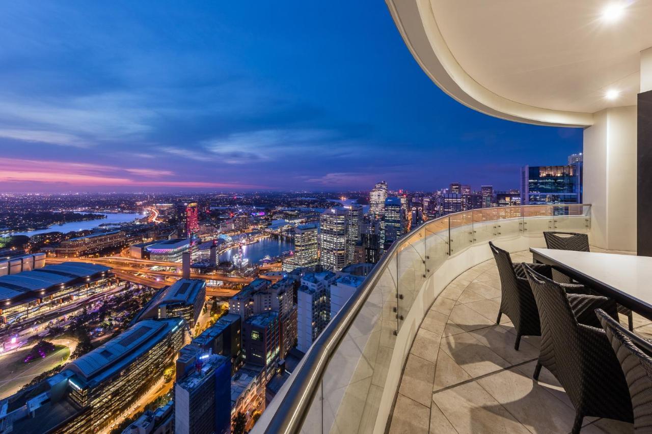 Meriton Suites Kent Street, Sydney Exterior photo The photo depicts a stunning cityscape view from a high balcony during twilight. The scene features a modern urban skyline, illuminated by city lights as night falls. You can see various buildings and structures, some with colorful lights. The balcon