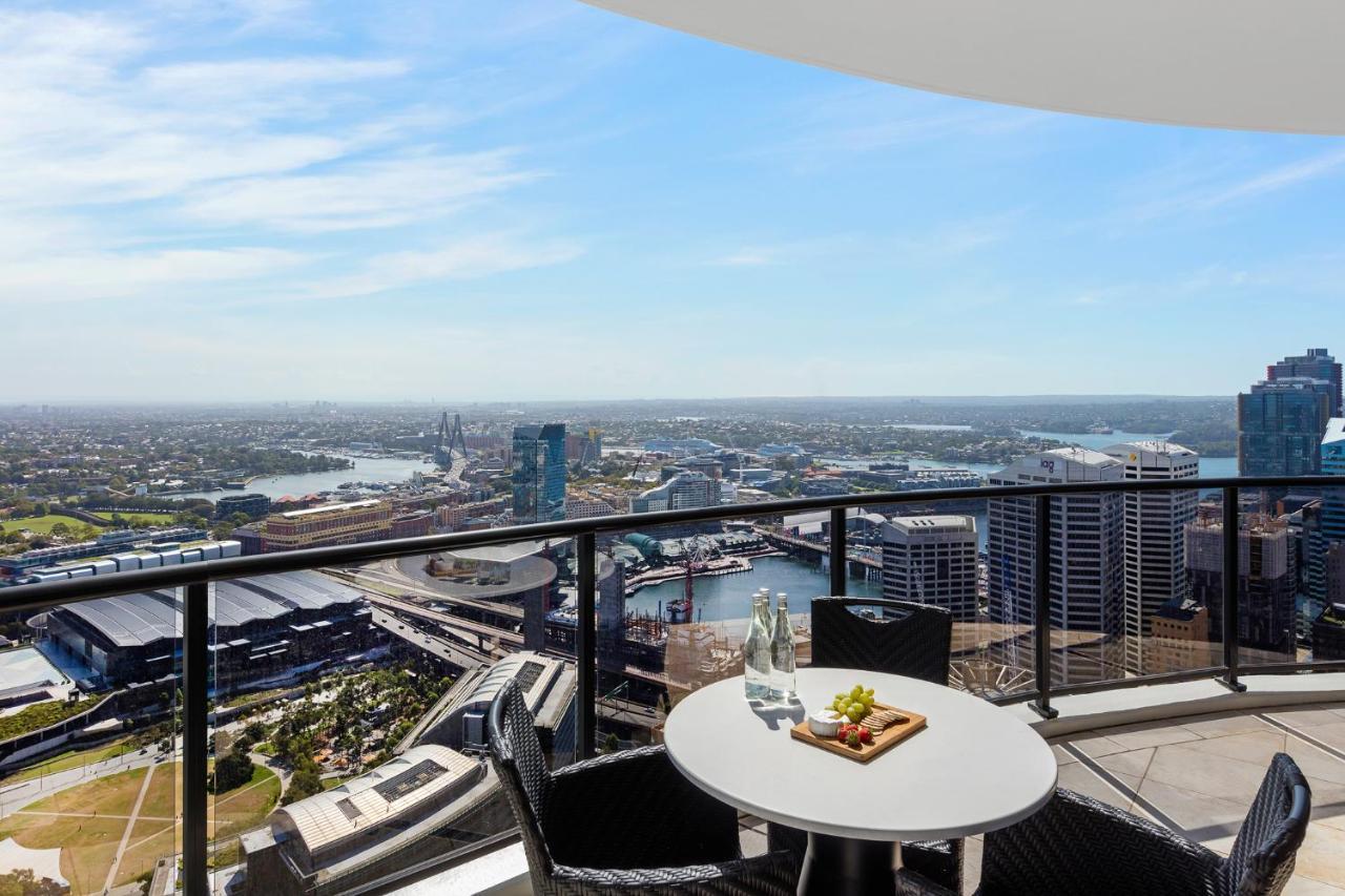 Meriton Suites Kent Street, Sydney Exterior photo The photo shows a balcony with a round table and two chairs. On the table, there is a small plate of fruit and a bottle of water. The view from the balcony is expansive, featuring a skyline with buildings, water, and distant land. The sky is clear wi