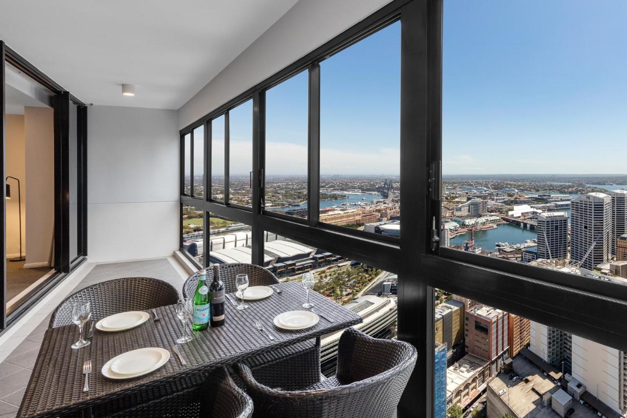 Meriton Suites Kent Street, Sydney Exterior photo The photo shows a balcony or terrace area of a high-rise building. There is a dining table set for four, with several plates and two wine glasses arranged on it. A bottle of wine or sparkling water is placed in the center of the table. The large wind