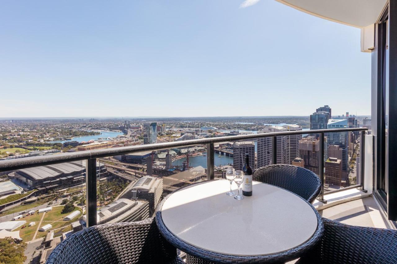 Meriton Suites Kent Street, Sydney Exterior photo The photo shows a balcony view from a high-rise building. There is a round table with two black wicker chairs, and a bottle of wine along with glasses placed on the table. The backdrop features a panoramic view of a cityscape, including rivers, build